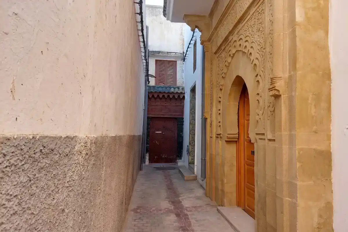 Vue d'une ruelle étroite dans la médina montrant une porte monumentale en pierre sculptée avec des arcs outrepassés typiques de l'art mauresque.