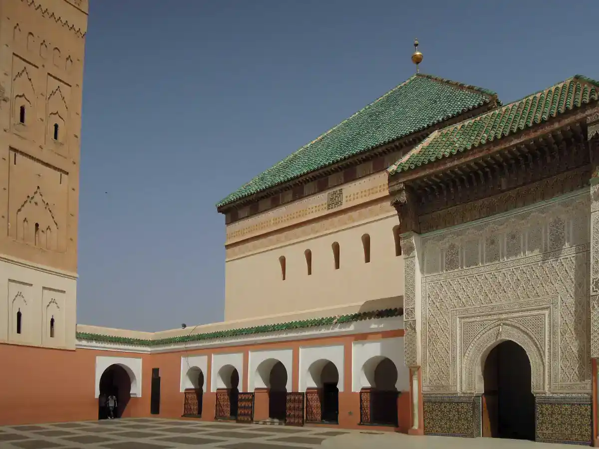 Sunlit courtyard of a mosque with orange walls, white arches, and a grand, intricately carved portal on the right.