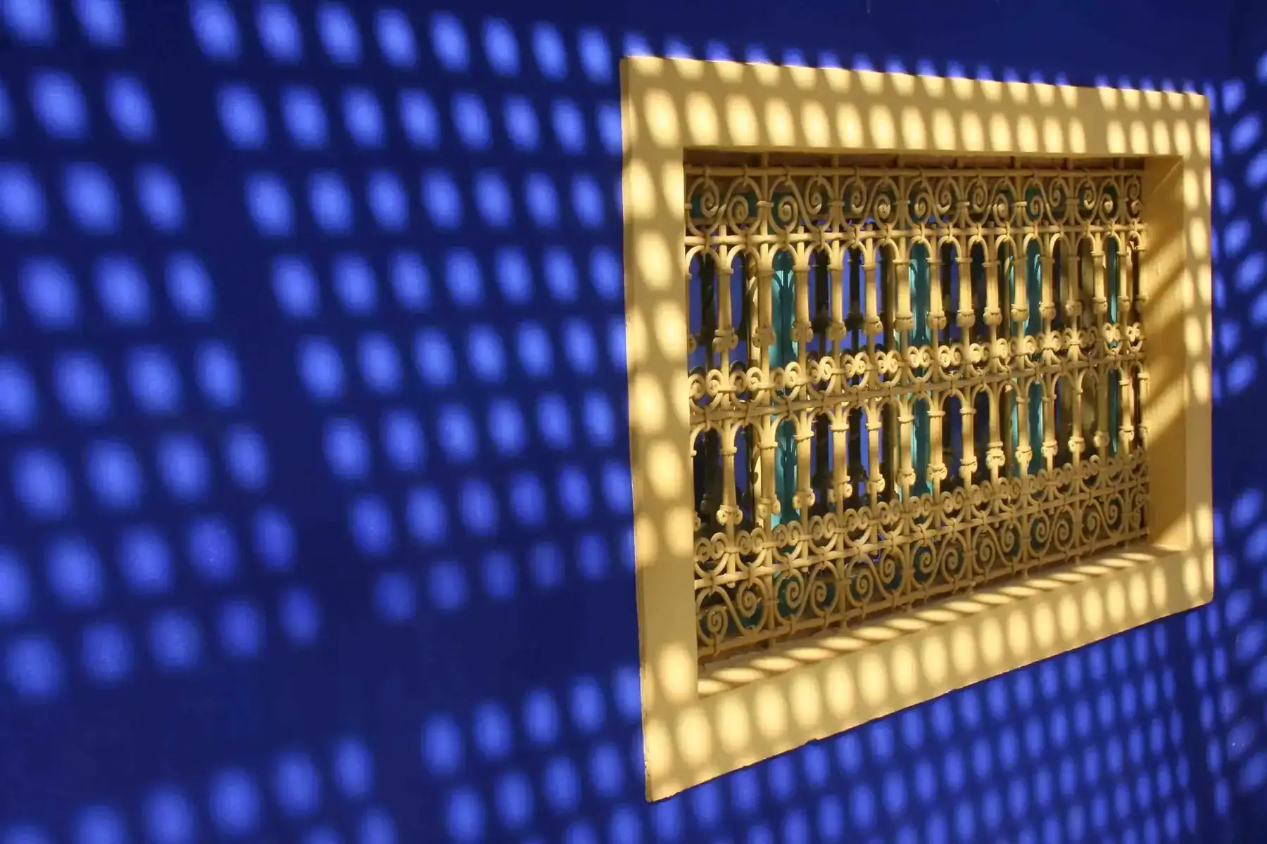 Blue wall with a yellow framed ornate iron grille window; dotted shadow pattern covers the wall created by the grille's shapes.