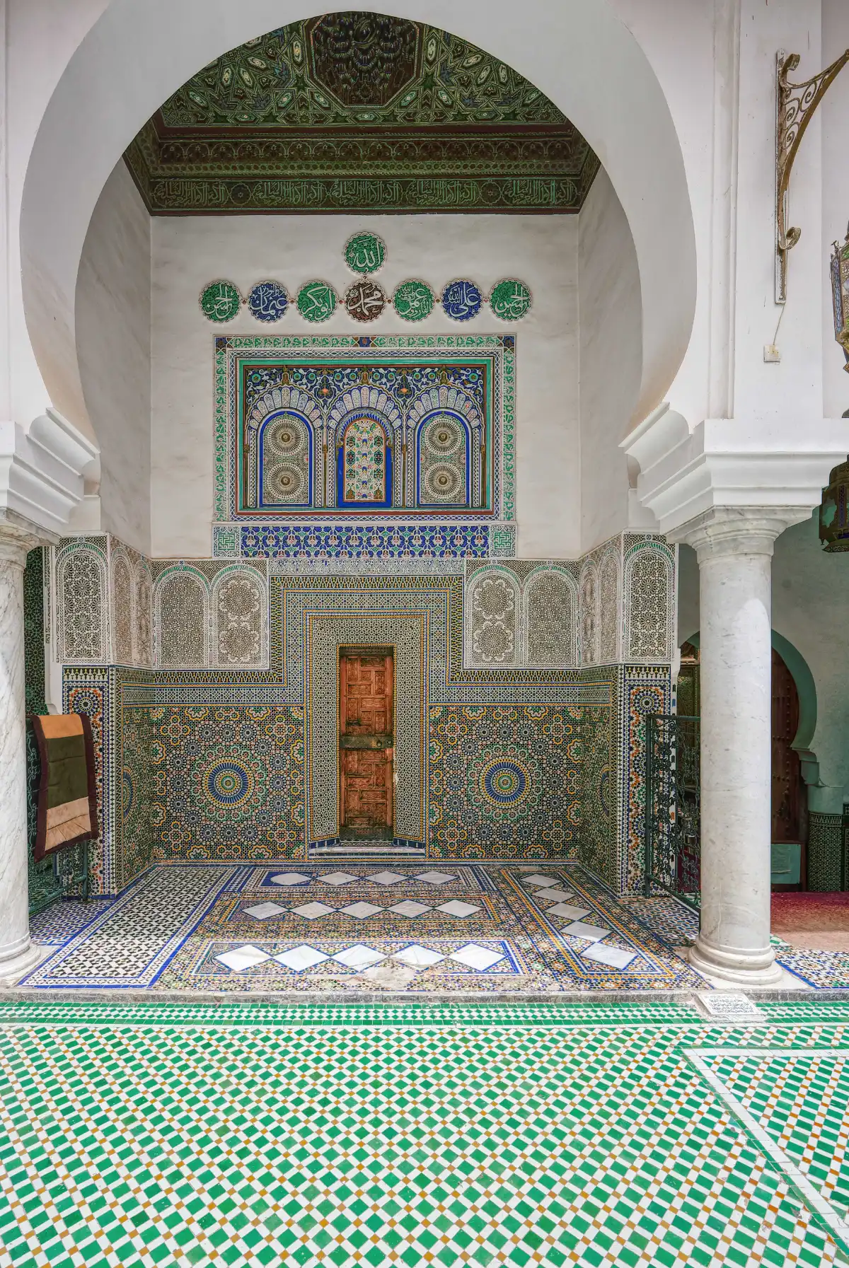 Patio intérieur de la mosquée Moulay Idriss à Fès avec murs en zelliges multicolores, colonnes en marbre blanc et calligraphie arabe ornementale.
