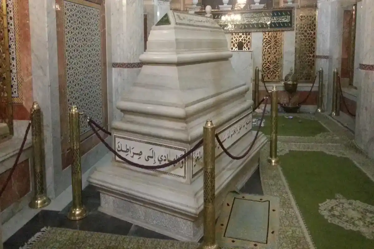 Marble mausoleum inside an ornate shrine, surrounded by gold stanchions and Arabic inscriptions on the base panel.