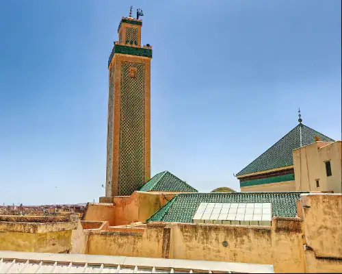 Vue sur le minaret de la zaouïa de Seïdina Ahmed Tijani à Fès, orné de zelliges verts, s'élevant au-dessus des toits de la médina sous un ciel bleu.
