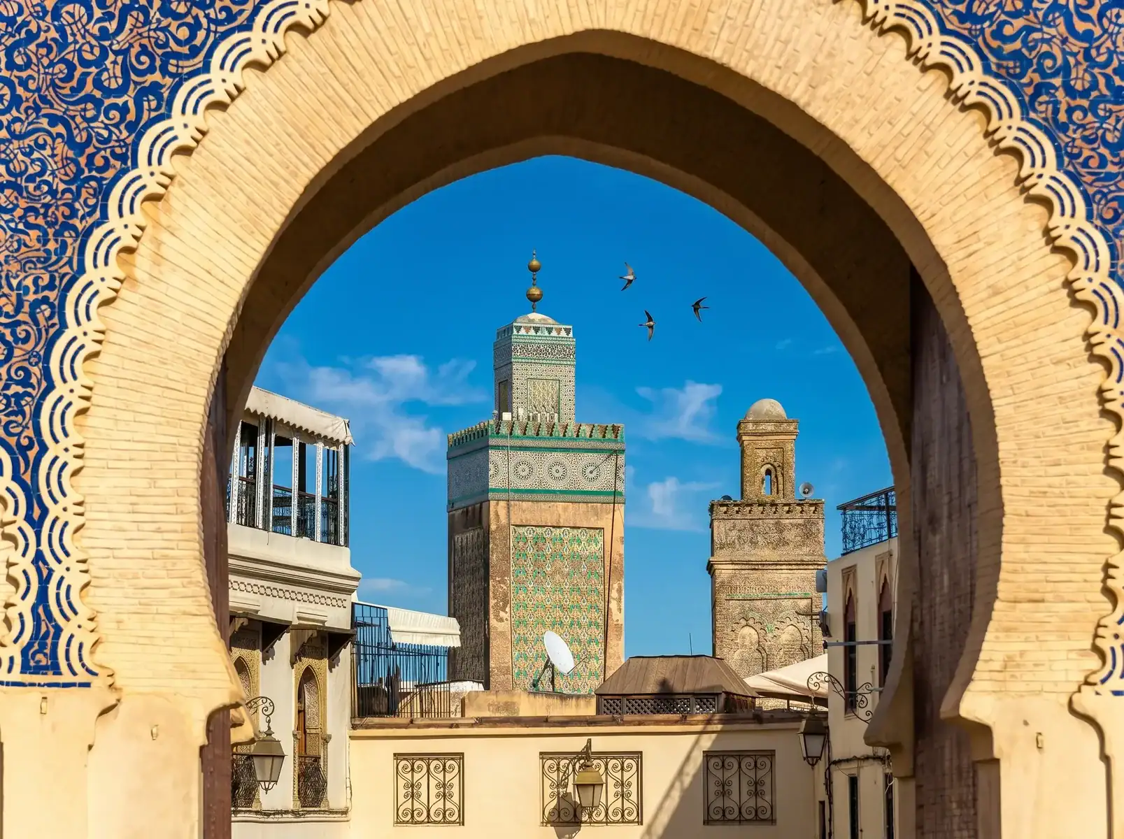 Archway with intricate beige bricks frames a turquoise-tiled minaret against a blue sky, with birds flying nearby.