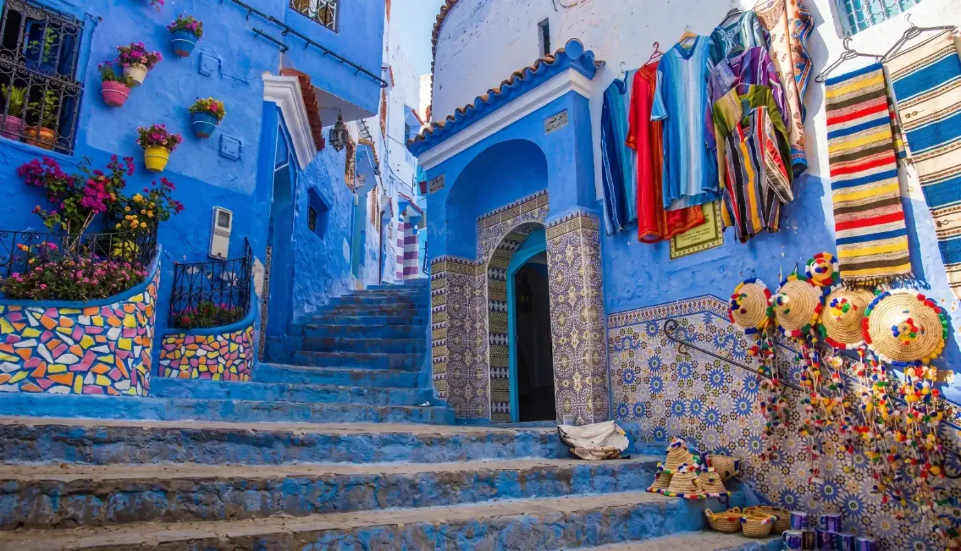 Entrée de la Zaouïa Ressounia dans une ruelle bleue typique de Chefchaouen, avec des mosaïques traditionnelles, des vêtements artisanaux suspendus et des chapeaux de paille colorés.