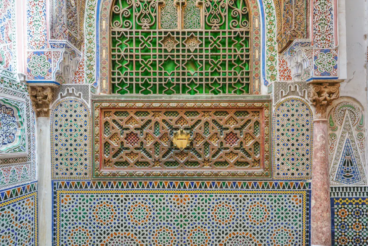 Détail architectural complexe de la mosquée de Moulay Idriss II à Fès au Maroc. L'image montre des mosaïques traditionnelles en zellige aux motifs géométriques colorés, surmontées de panneaux en bois richement sculptés abritant une fente à aumône en laiton, et d'une grille traditionnelle verte, encadrés par des sculptures en stuc et une colonne en marbre.